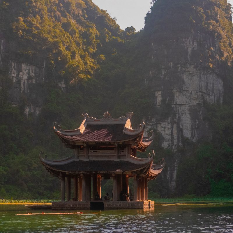 A wooden pagoda standing on a calm lake below limestone cliffs in Ninh B&igrave;nh, Vietnam.